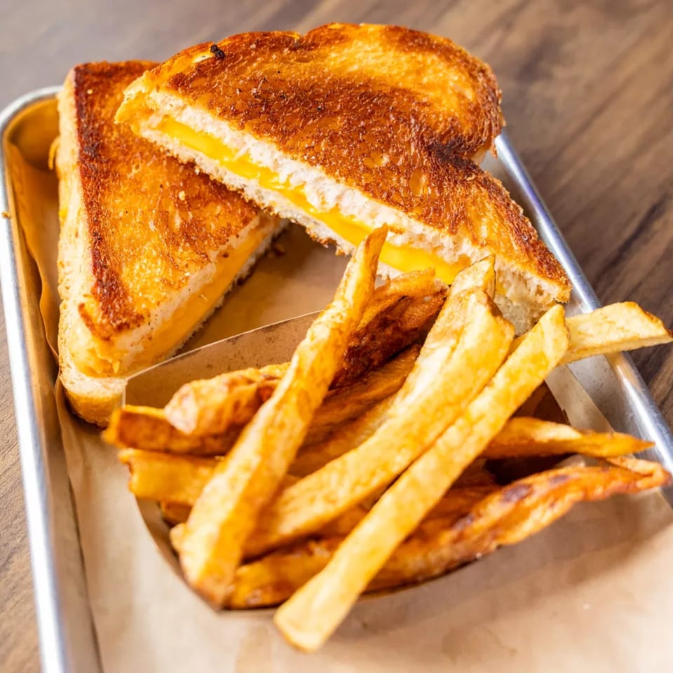 Golden grilled cheese sandwich cut in half, revealing melted cheese, served with crispy fries on a metal tray.