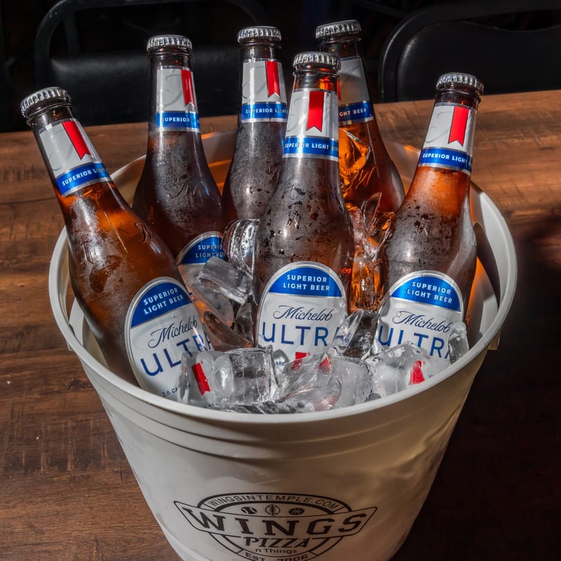 Six Michelob Ultra beer bottles in a white bucket filled with ice on a wooden table.