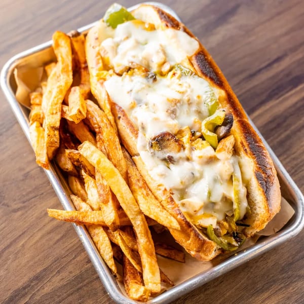 Cheesesteak sandwich with melted cheese, peppers, and mushrooms, served with fries on a metal tray.