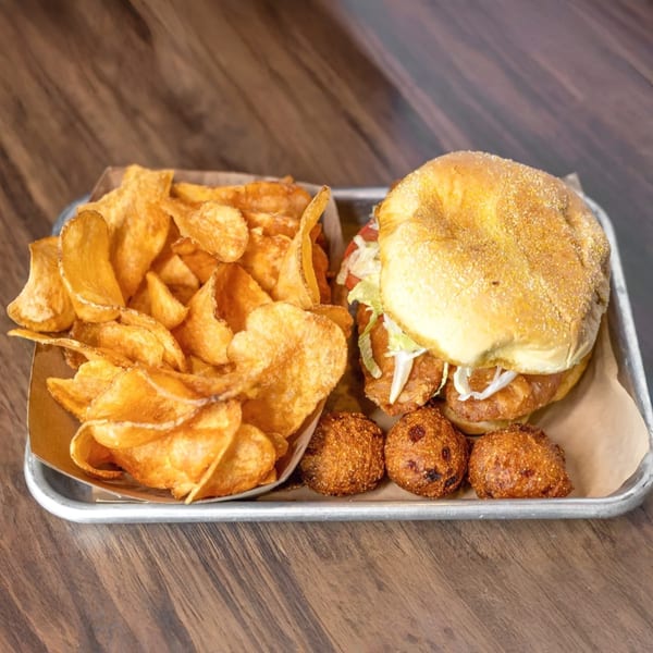 A fried chicken sandwich, kettle chips, and hush puppies on a metal tray.