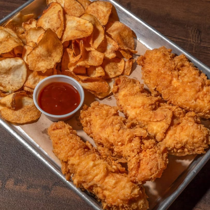 Crispy fried chicken tenders, a side of golden potato chips, and a cup of dipping sauce on a metal tray.