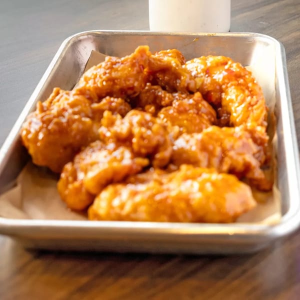 A metal tray filled with golden-brown, saucy boneless chicken wings, with a white dipping sauce in the background.